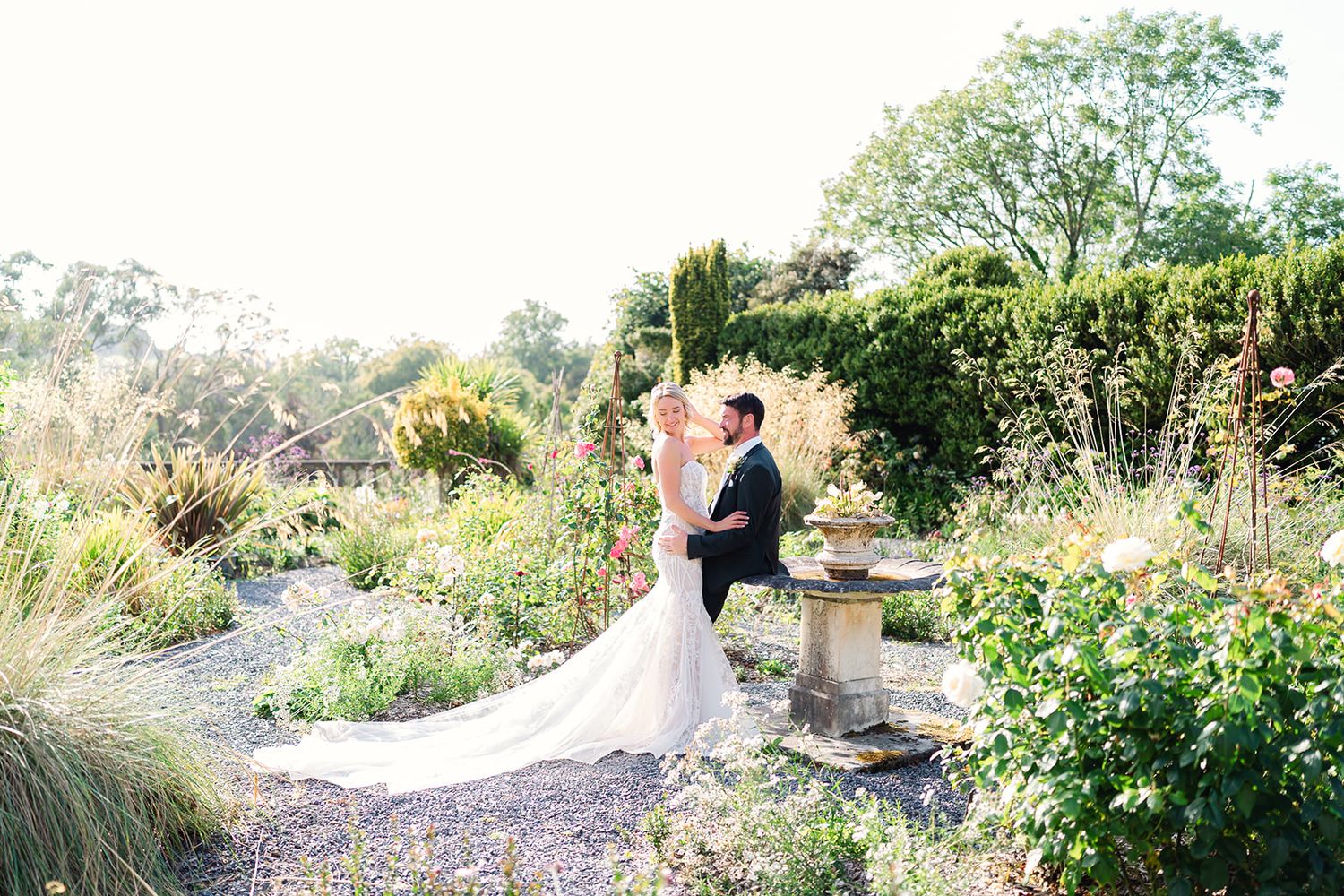A wedding couple embraces in a lush garden setting with flowing dress and natural scenery.
