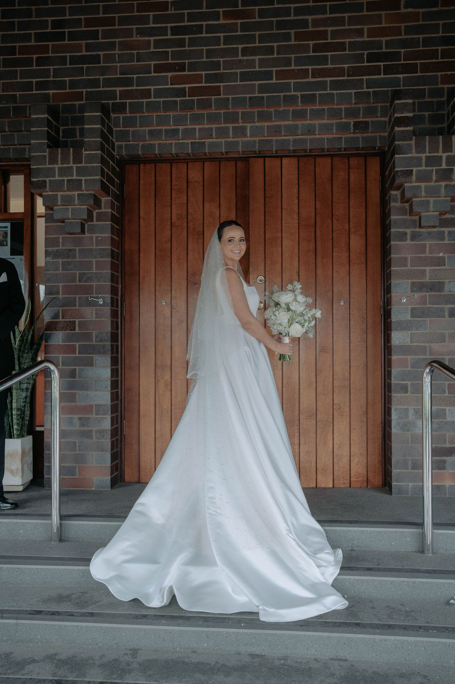 A bride in an elegant white satin wedding gown with a flowing train poses by a wooden door against dark brick walls.