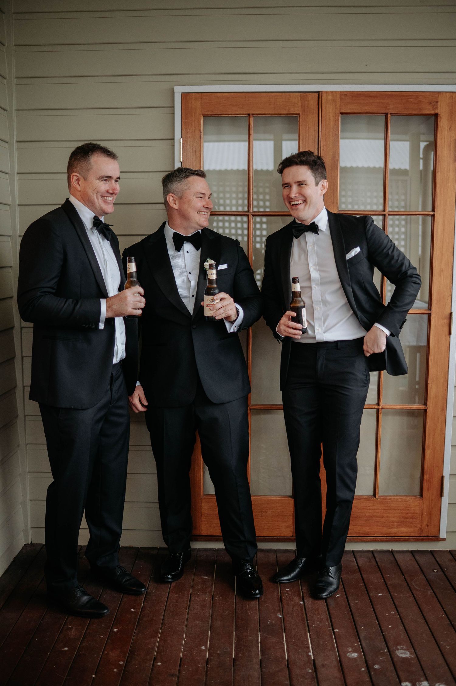 Three people in black tuxedos sharing drinks on a wooden deck during a formal event.