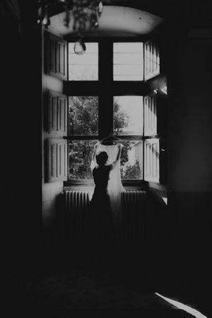 A silhouette against a bright window opening curtains in a moody black and white photograph.