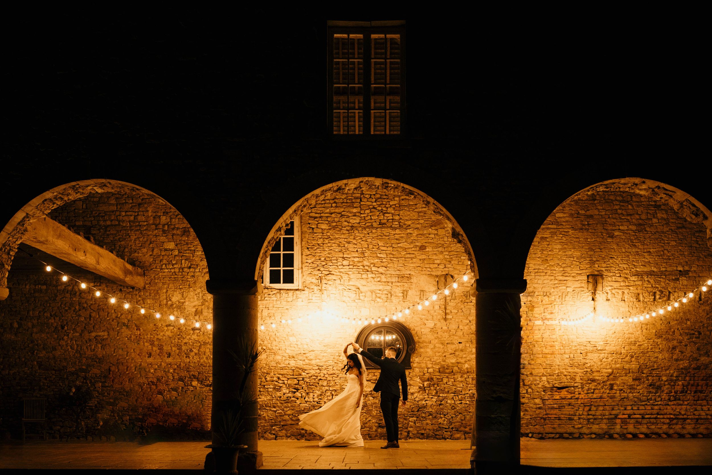 A romantic dance silhouette under three stone archways with warm lighting and string lights.