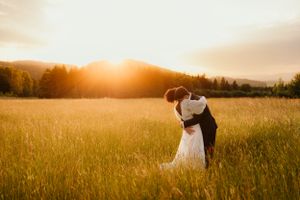 Couple embracing in sunlit meadow with mountains in background during sunset.