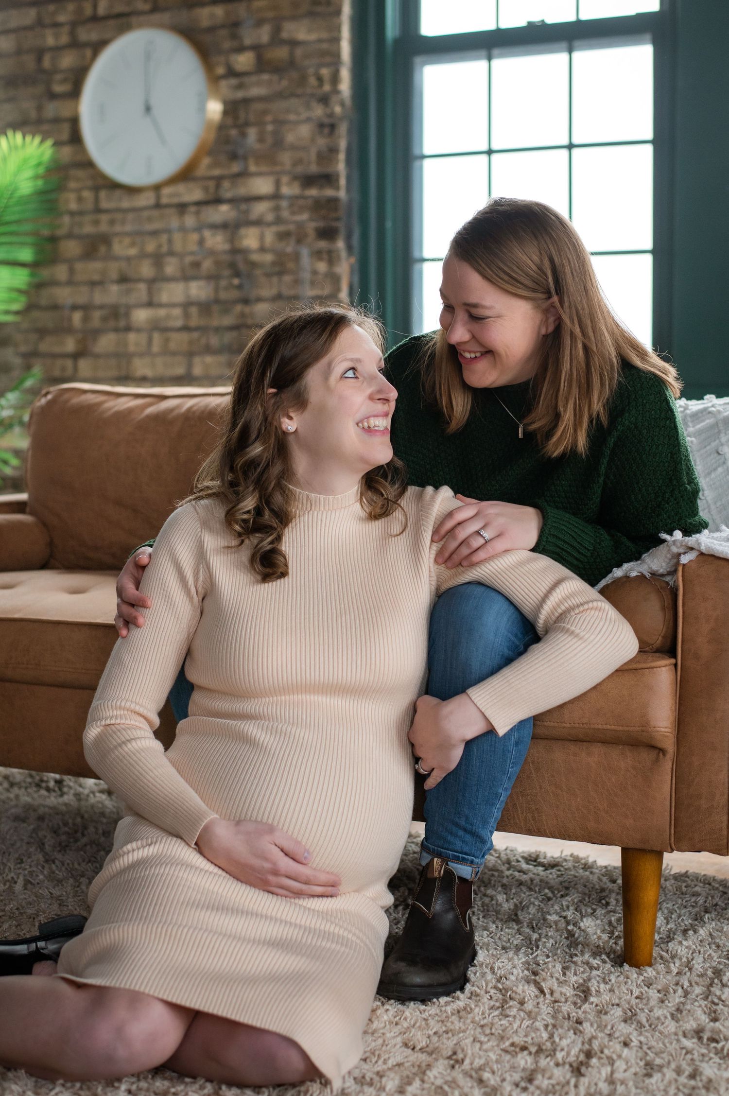 Two people share a warm moment on a brown couch, with one wearing a cream dress showing a baby bump, in a cozy room.