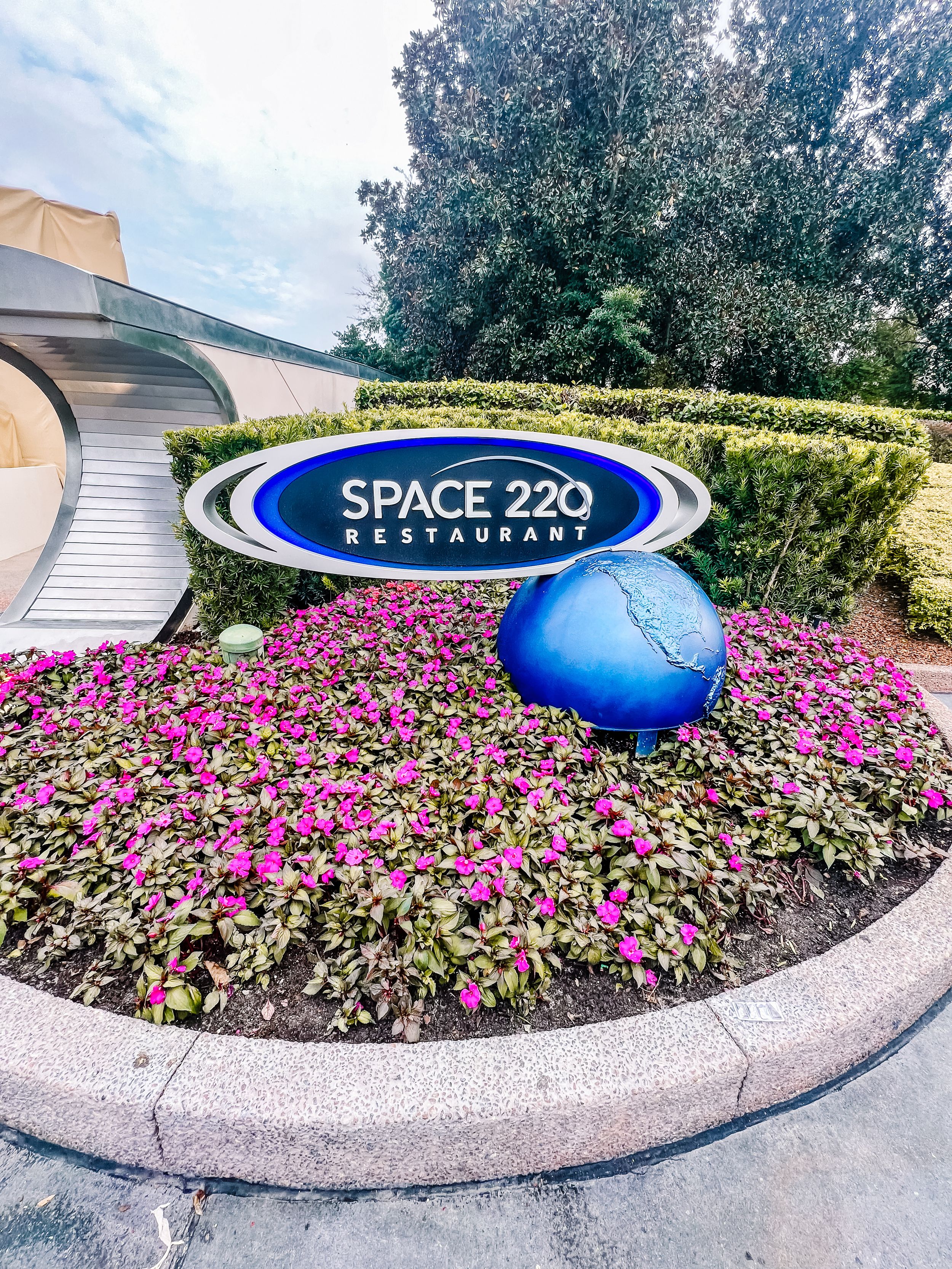 Space 220 restaurant sign surrounded by pink flowers and a blue sphere in a landscaped garden.