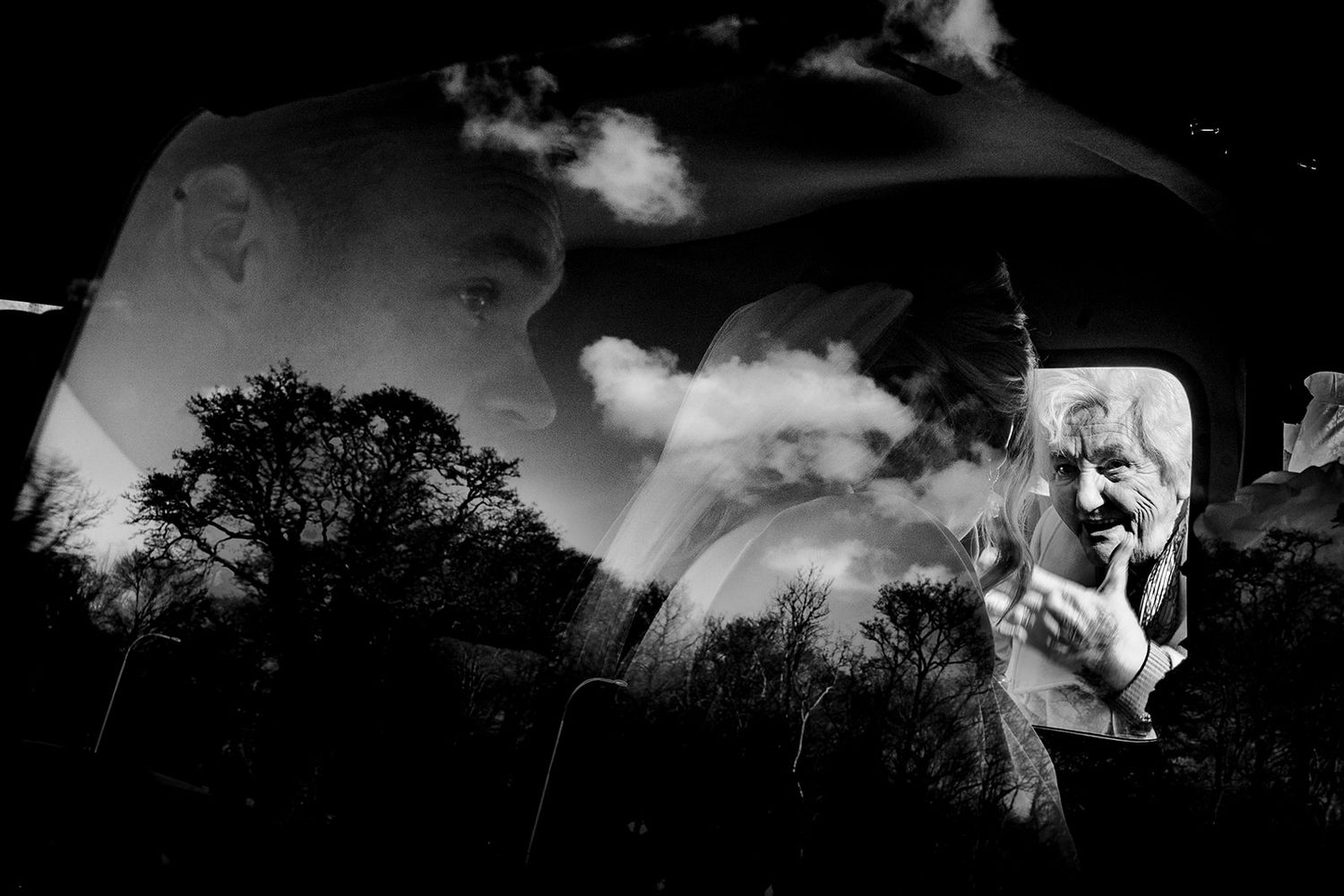 A dramatic black and white double exposure photograph combining silhouetted trees with ethereal clouds and fabric.