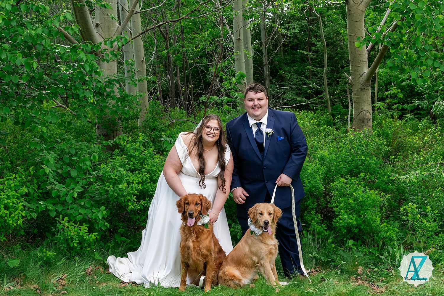 A couple in wedding attire poses with their two brown dogs against a lush green forest backdrop.