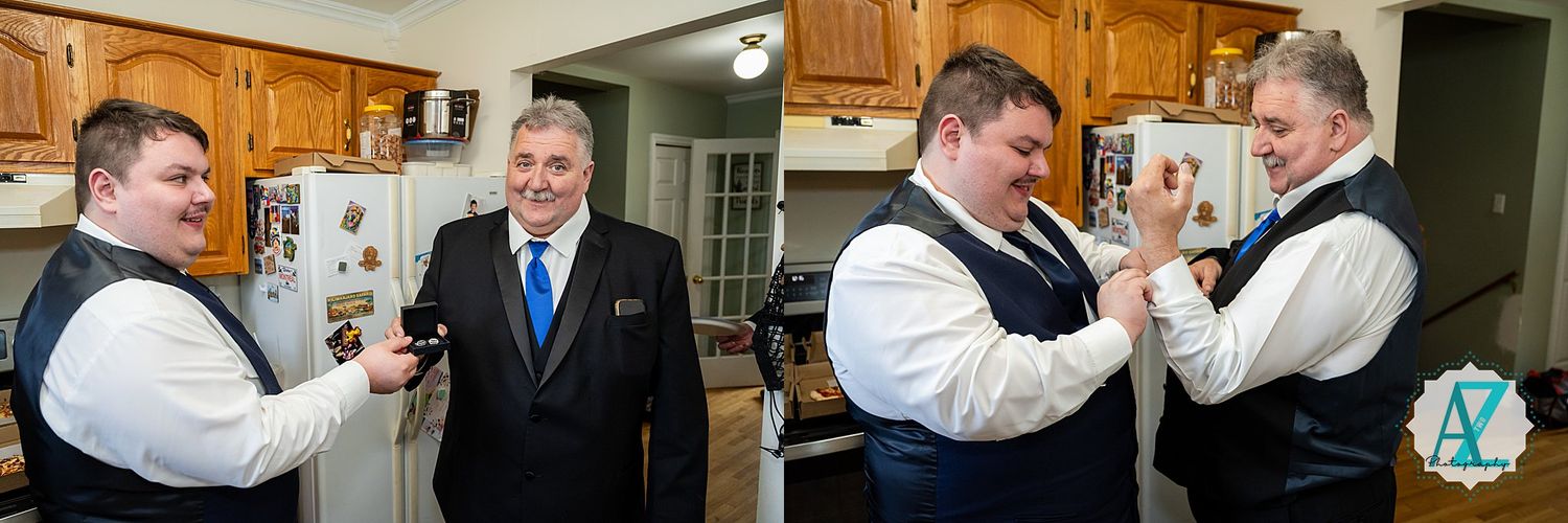 Several people in formal attire interact in pairs during an indoor gathering with wooden kitchen cabinets visible.