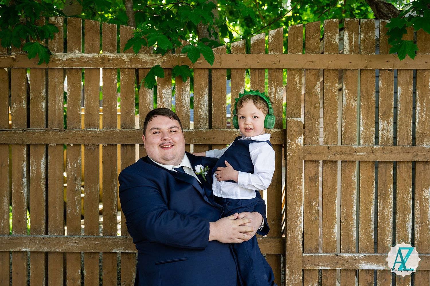 Two people stand together against a wooden fence in a backyard with green foliage overhead.