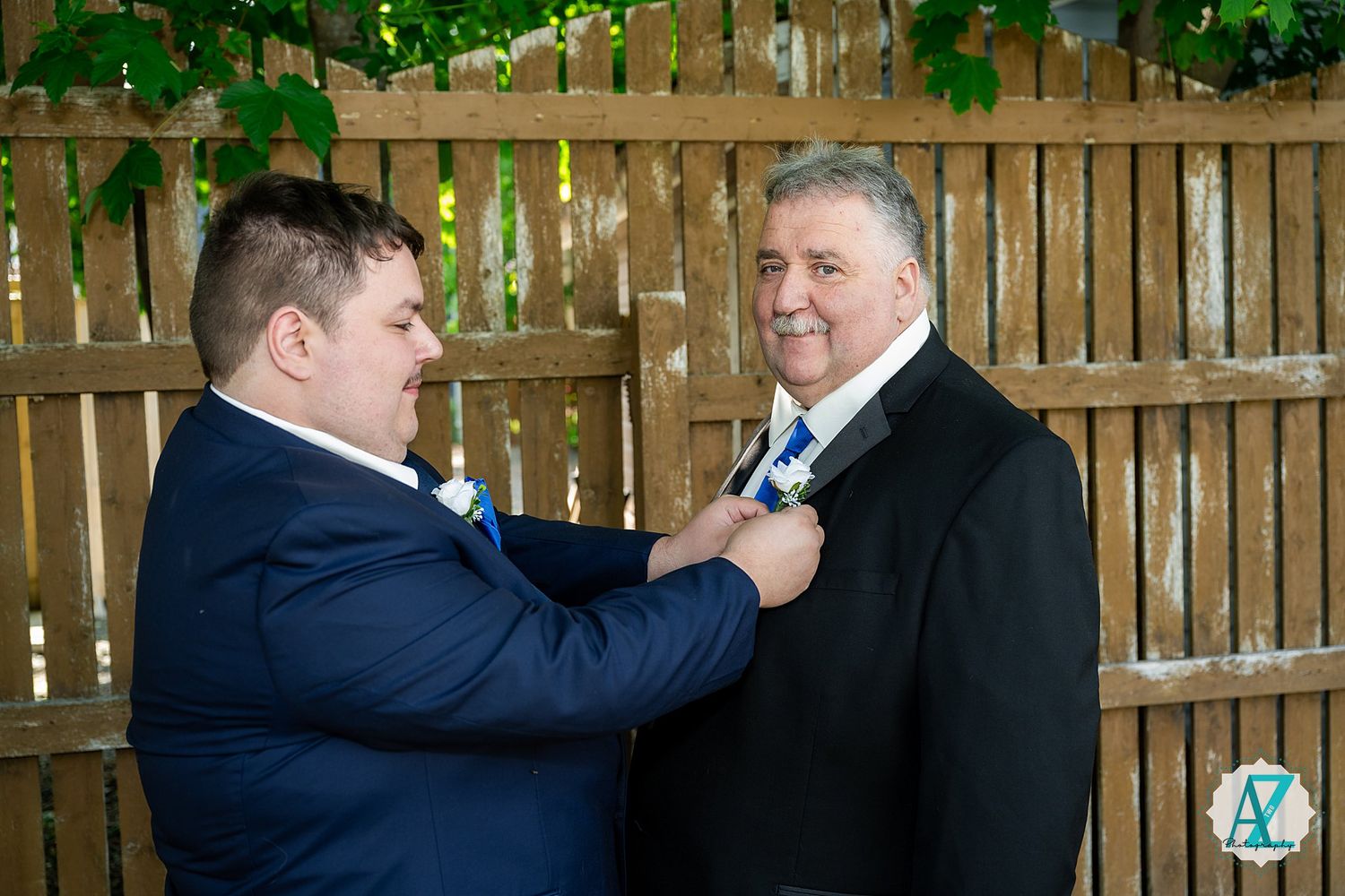 Two people in navy and black suits help each other adjust their ties while standing by a wooden fence outdoors.