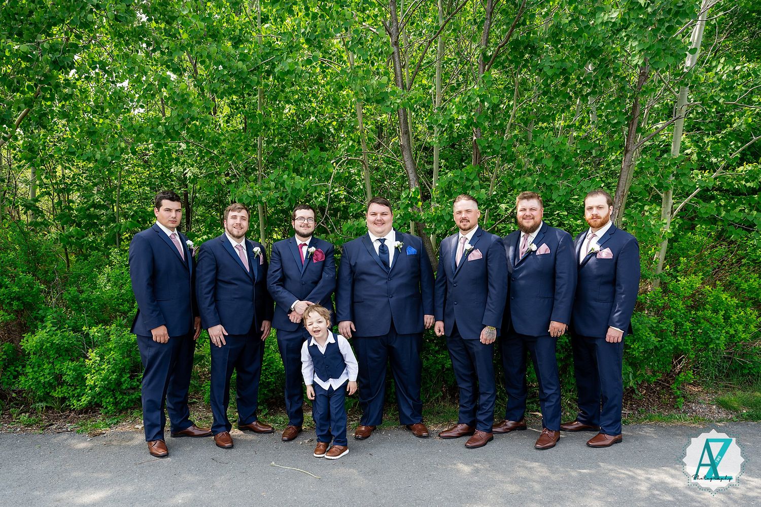 A group of men in matching navy suits stand in a row outdoors against a green forest backdrop.