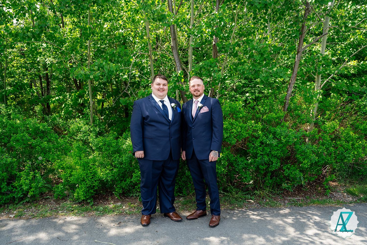 Two people in navy suits and brown dress shoes stand together on a path with lush green foliage in the background.