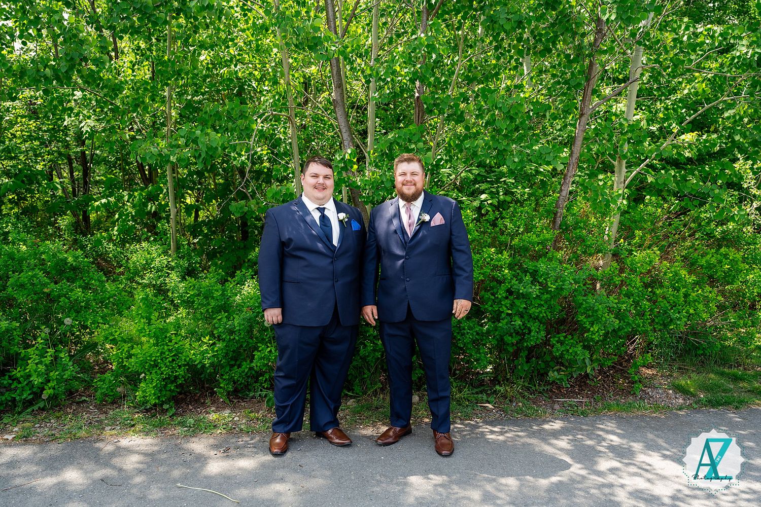 Two people in navy suits stand together on a pathway with lush green foliage in the background.