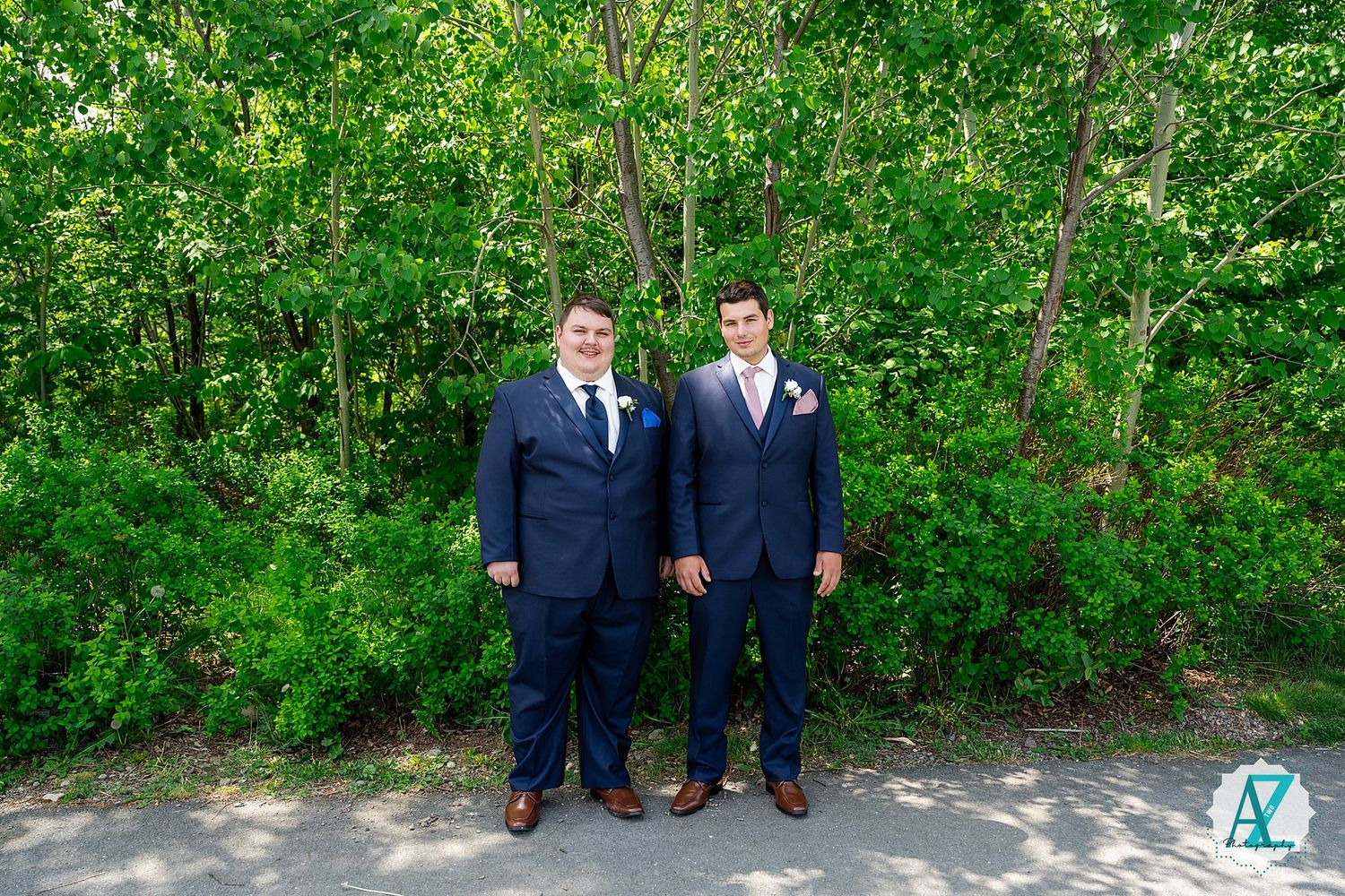 Groomsmen in navy blue suits standing outdoors along a lush green hedge on a sunny day.