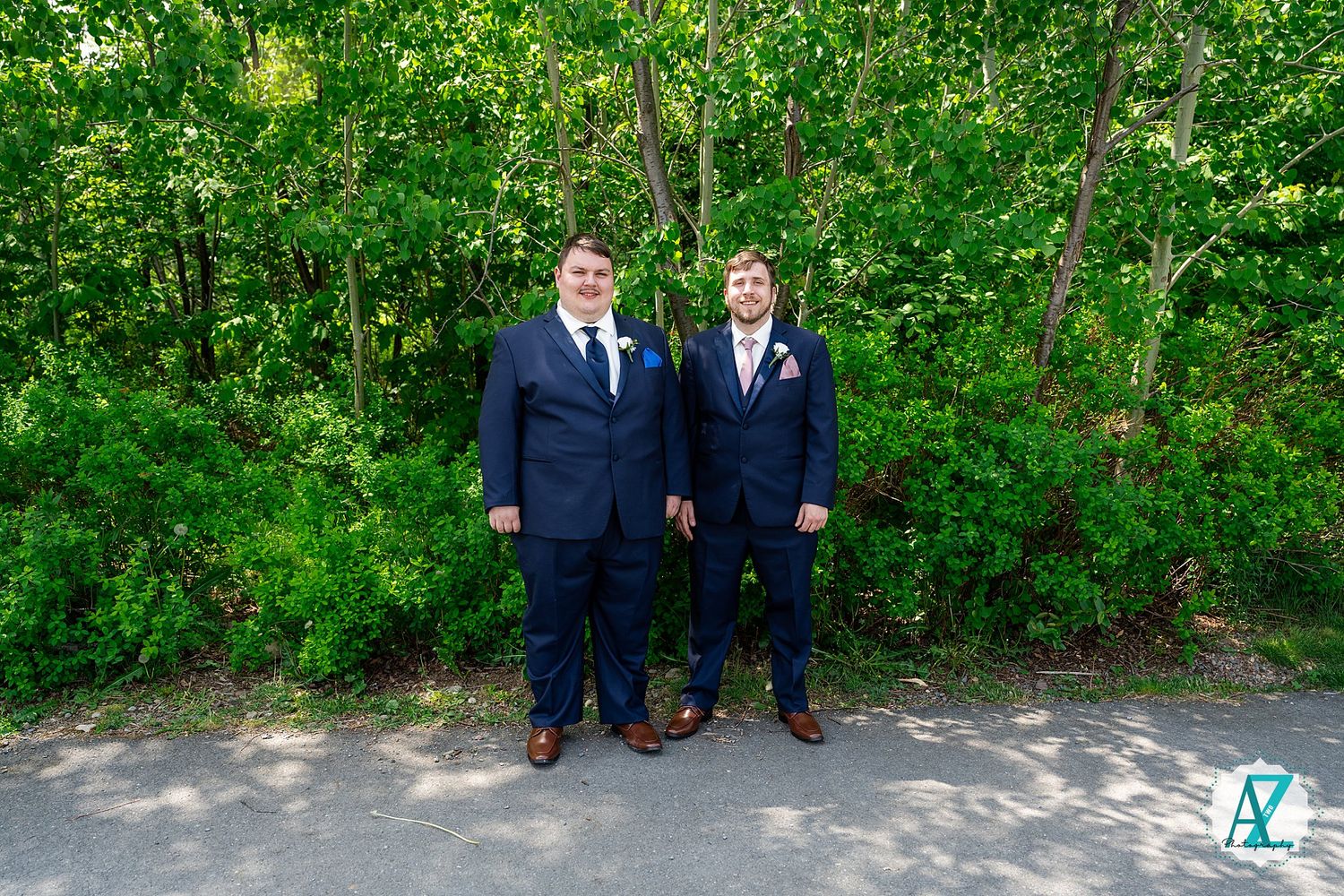 Two people wearing navy suits and ties stand together on a path with lush green foliage in the background.