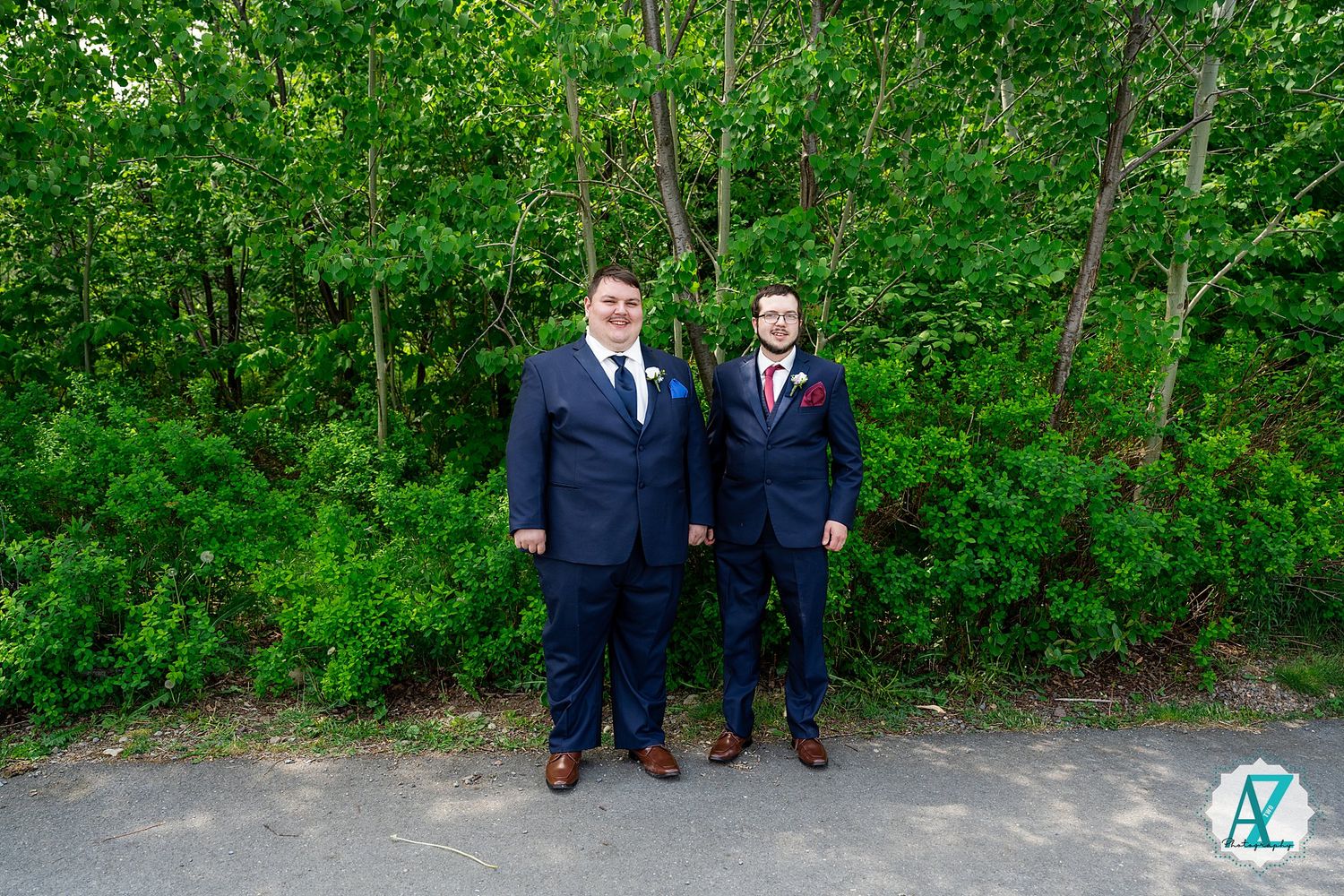 Two people in navy suits stand together on a path with lush green foliage in the background.