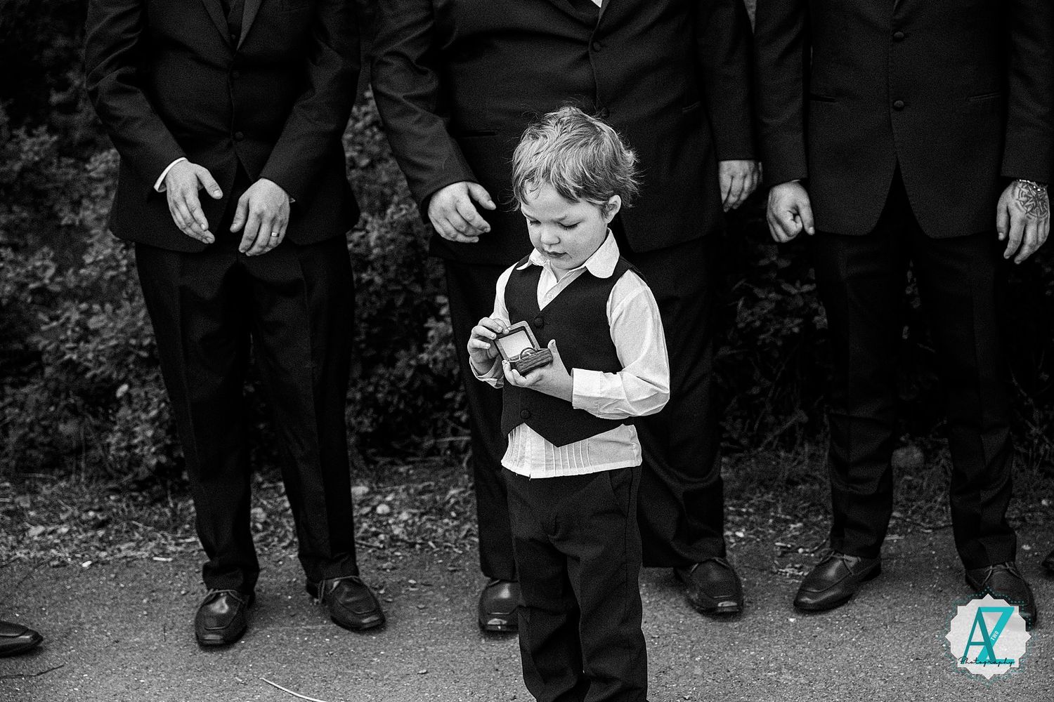 Black and white photo of a young ring bearer in formal attire playing with an object while groomsmen stand behind him.