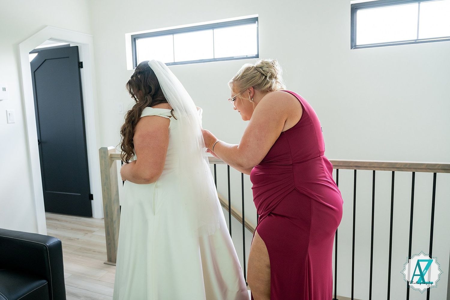 A person in a burgundy dress helps adjust a white wedding gown in a brightly lit room.