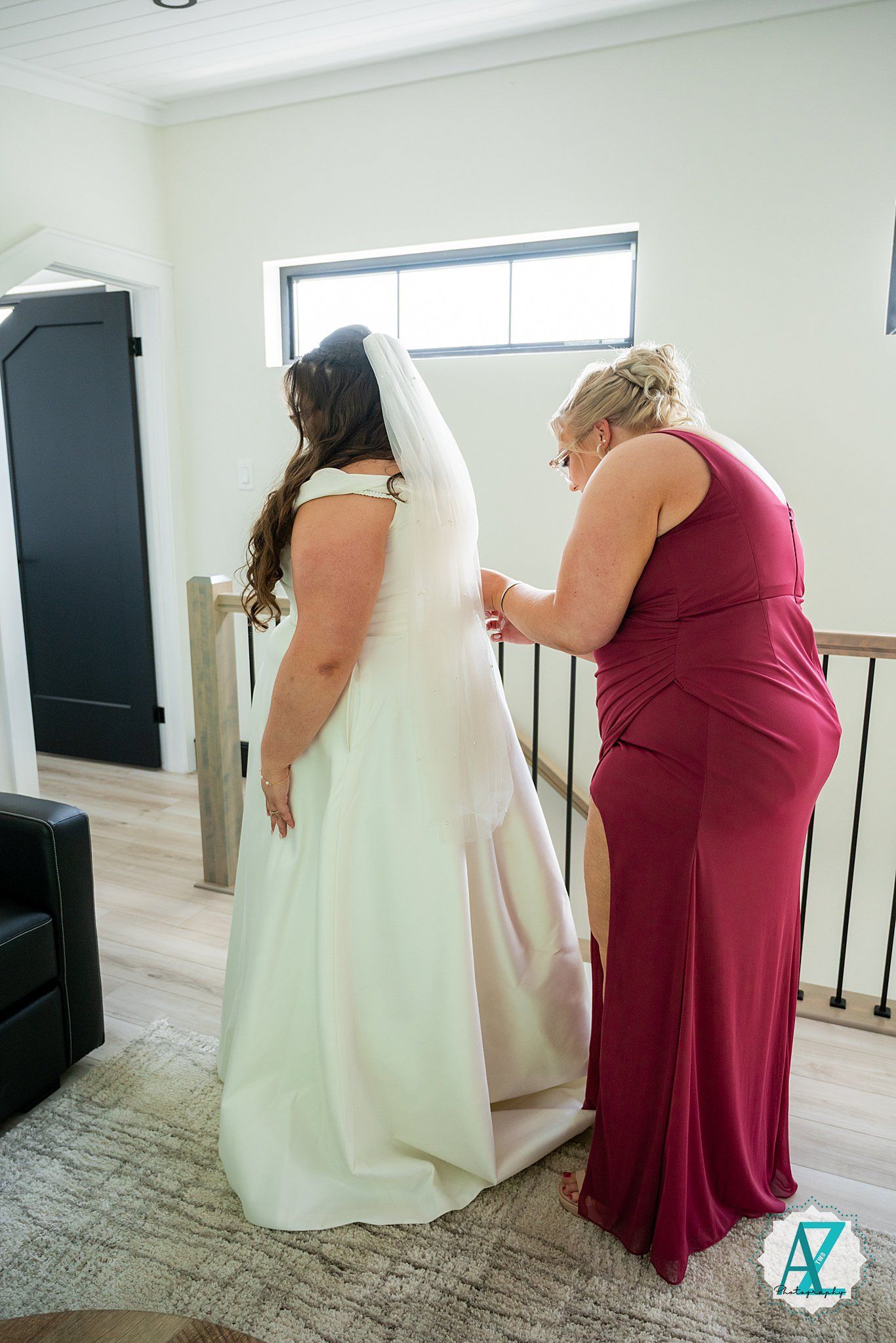 A person in a white wedding dress being helped with final preparations by an attendant in a burgundy gown.
