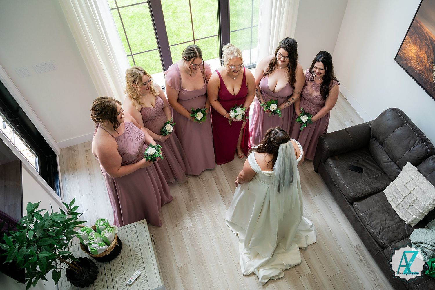 Bridesmaids in dusty rose dresses gather by bright windows in a white room during wedding preparations.