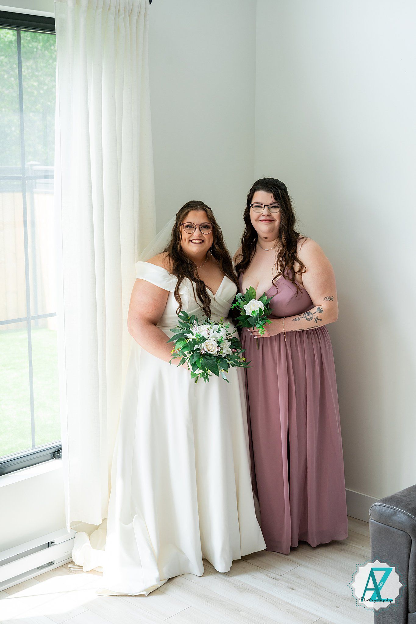 Two figures in wedding attire pose by window, wearing white gown and mauve bridesmaid dress with floral bouquet.