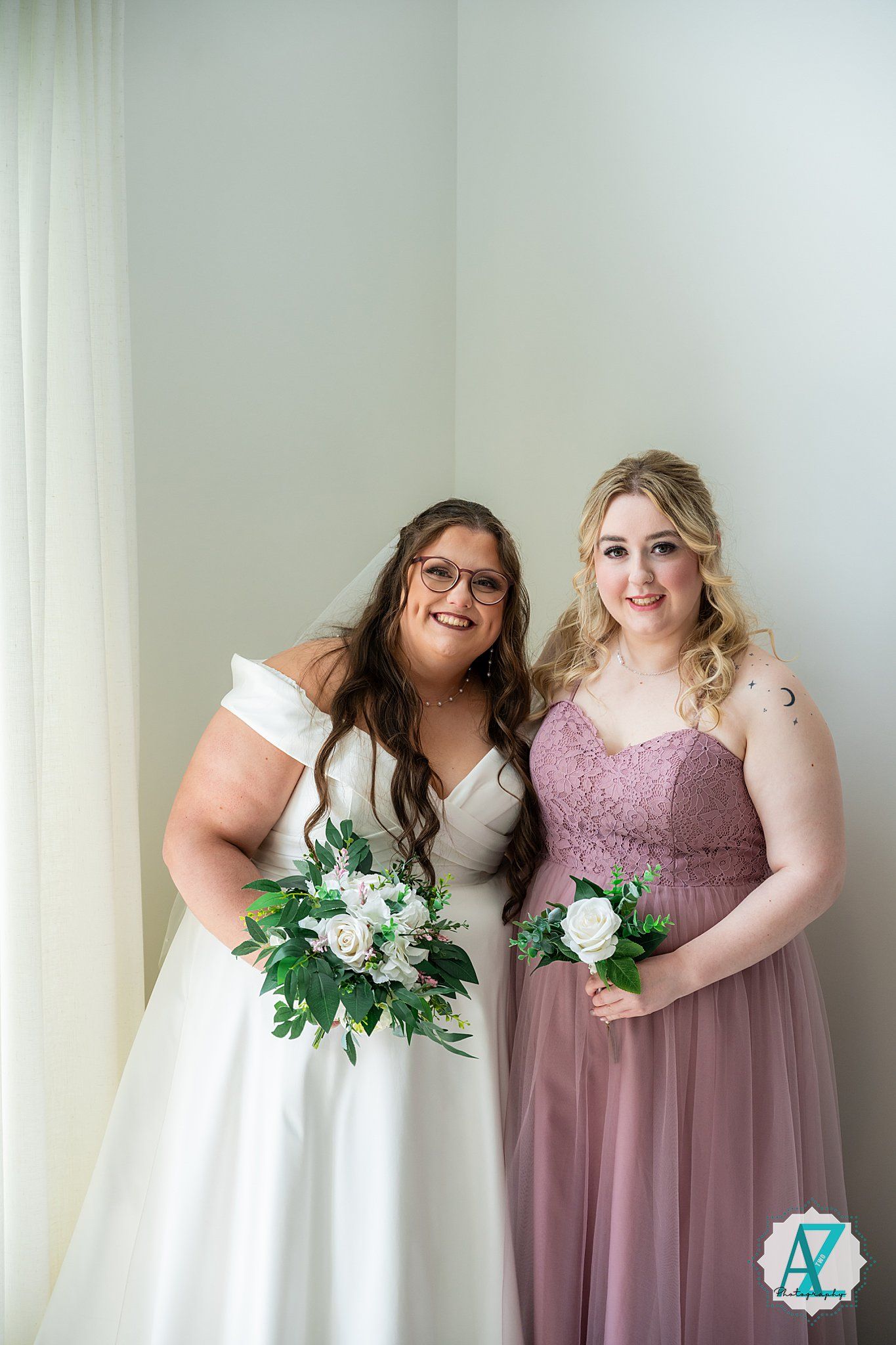 Two people in wedding attire pose together holding white and green floral bouquets.