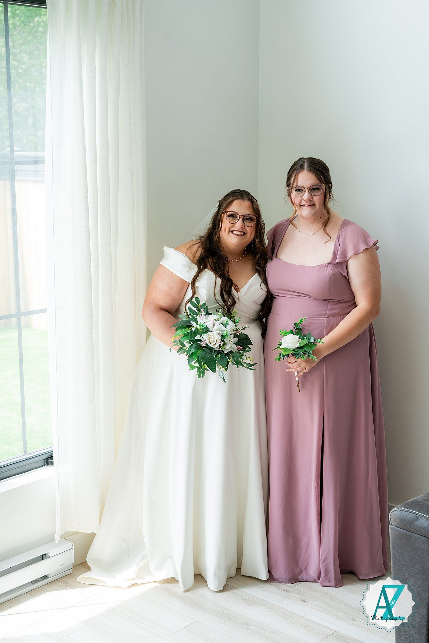 Wedding party members in pink dresses stand near sunlit windows holding white and green bouquets.