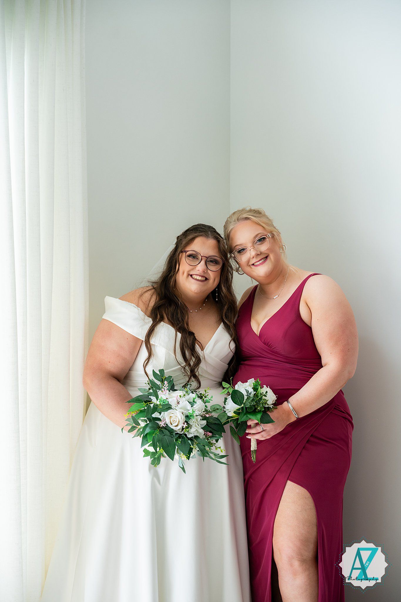 Two figures in burgundy and white dresses stand together holding floral bouquets.