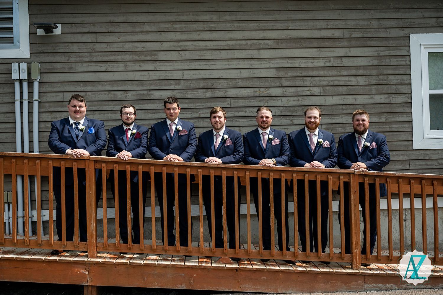 A group of groomsmen in matching navy suits stand together on a wooden deck railing during a wedding.