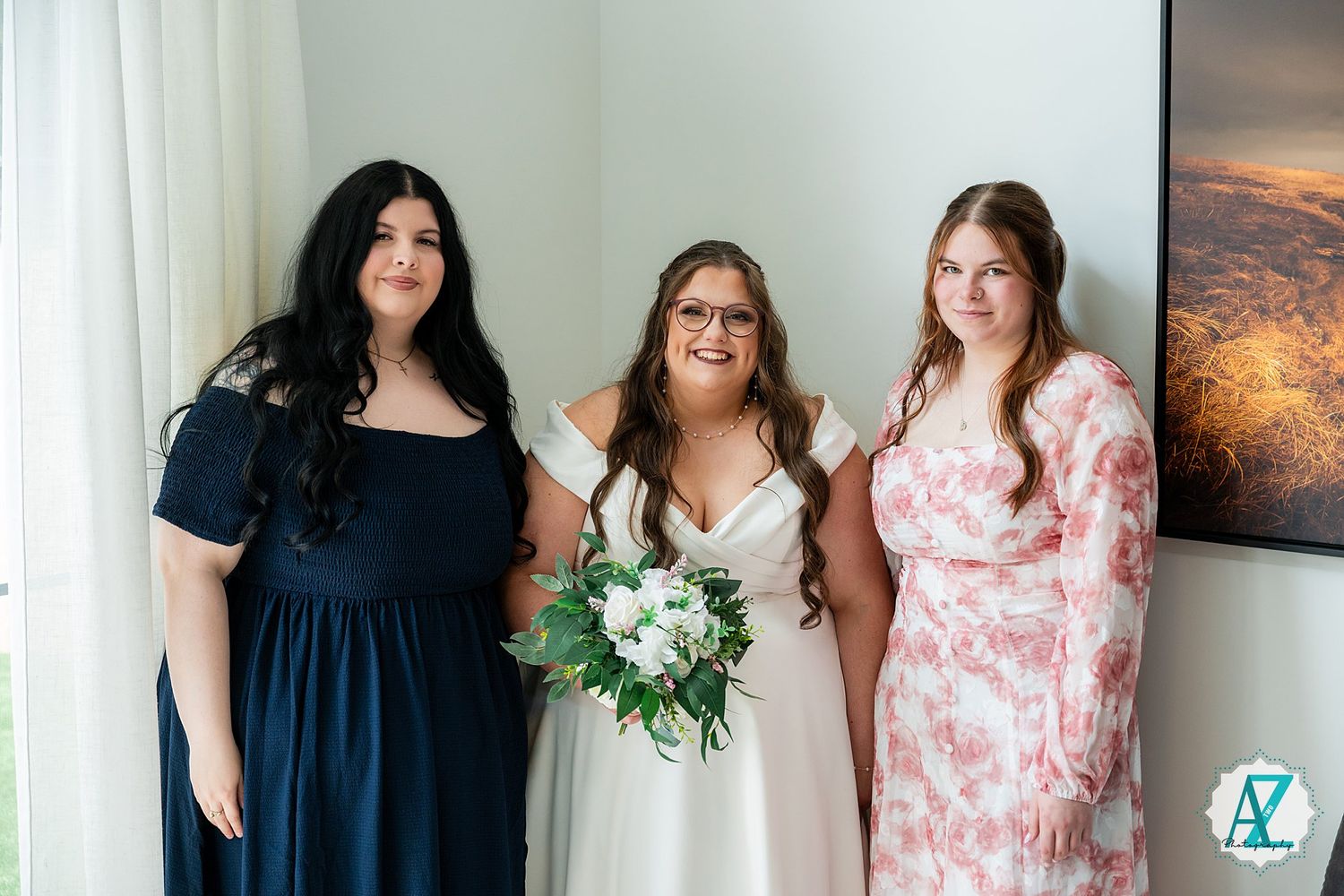 Three women pose together at a wedding celebration with floral bouquet and nature artwork in background.