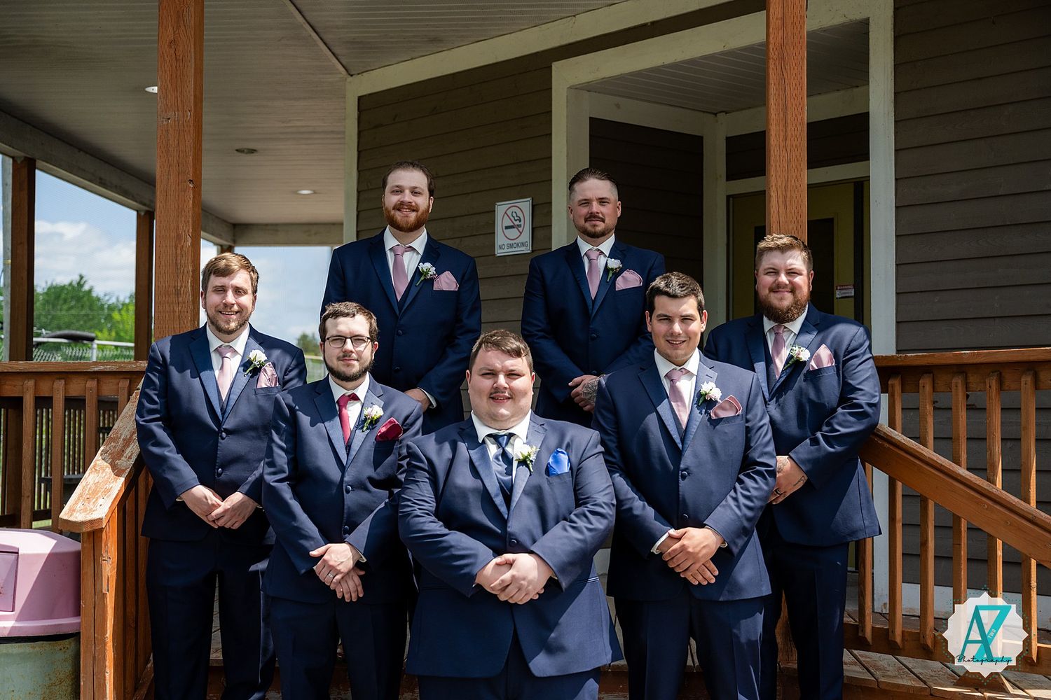 A group of groomsmen wearing navy suits pose together on a wooden front porch of a house.