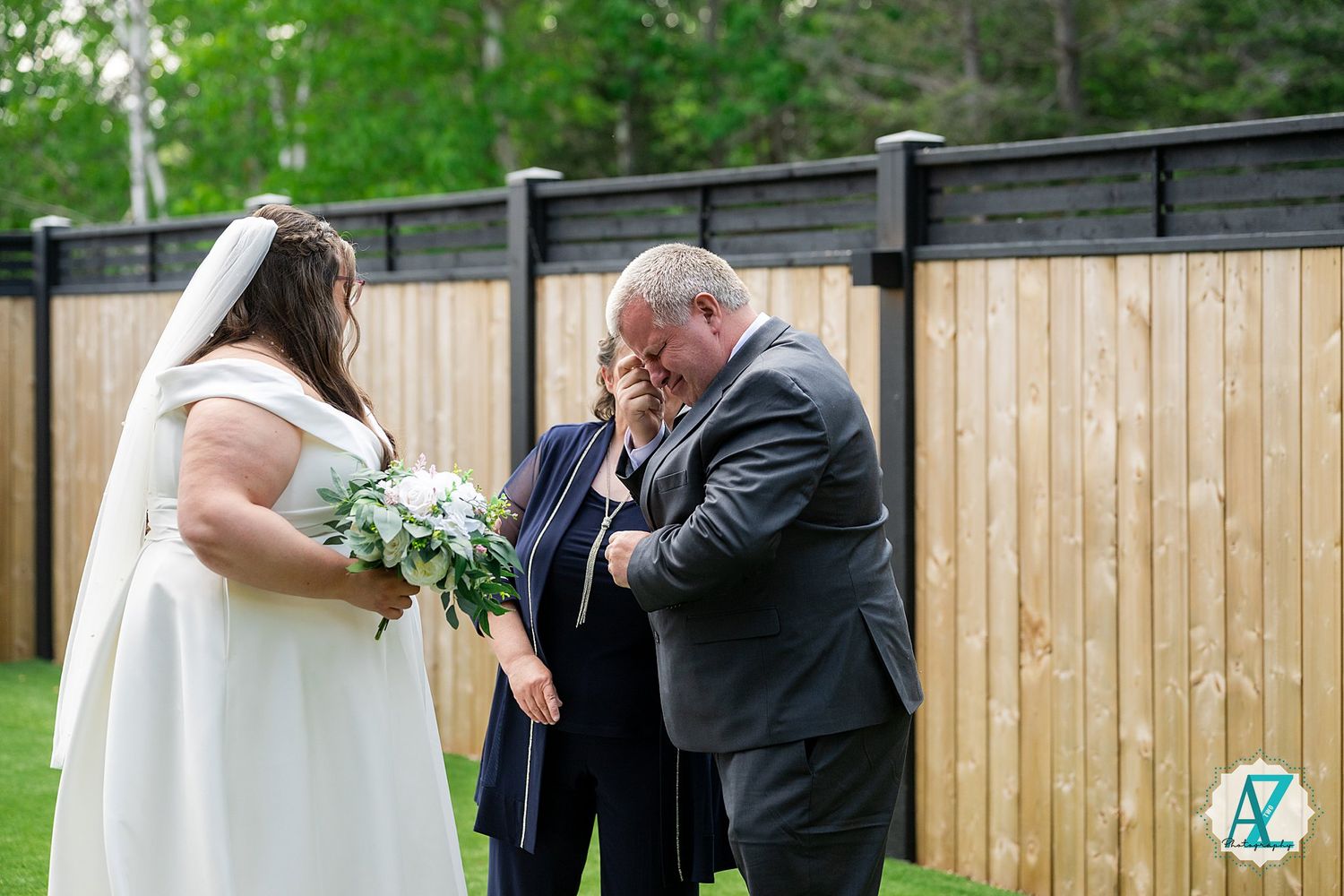 An emotional backyard garden wedding ceremony scene with a white wooden fence and lush green foliage in the background.