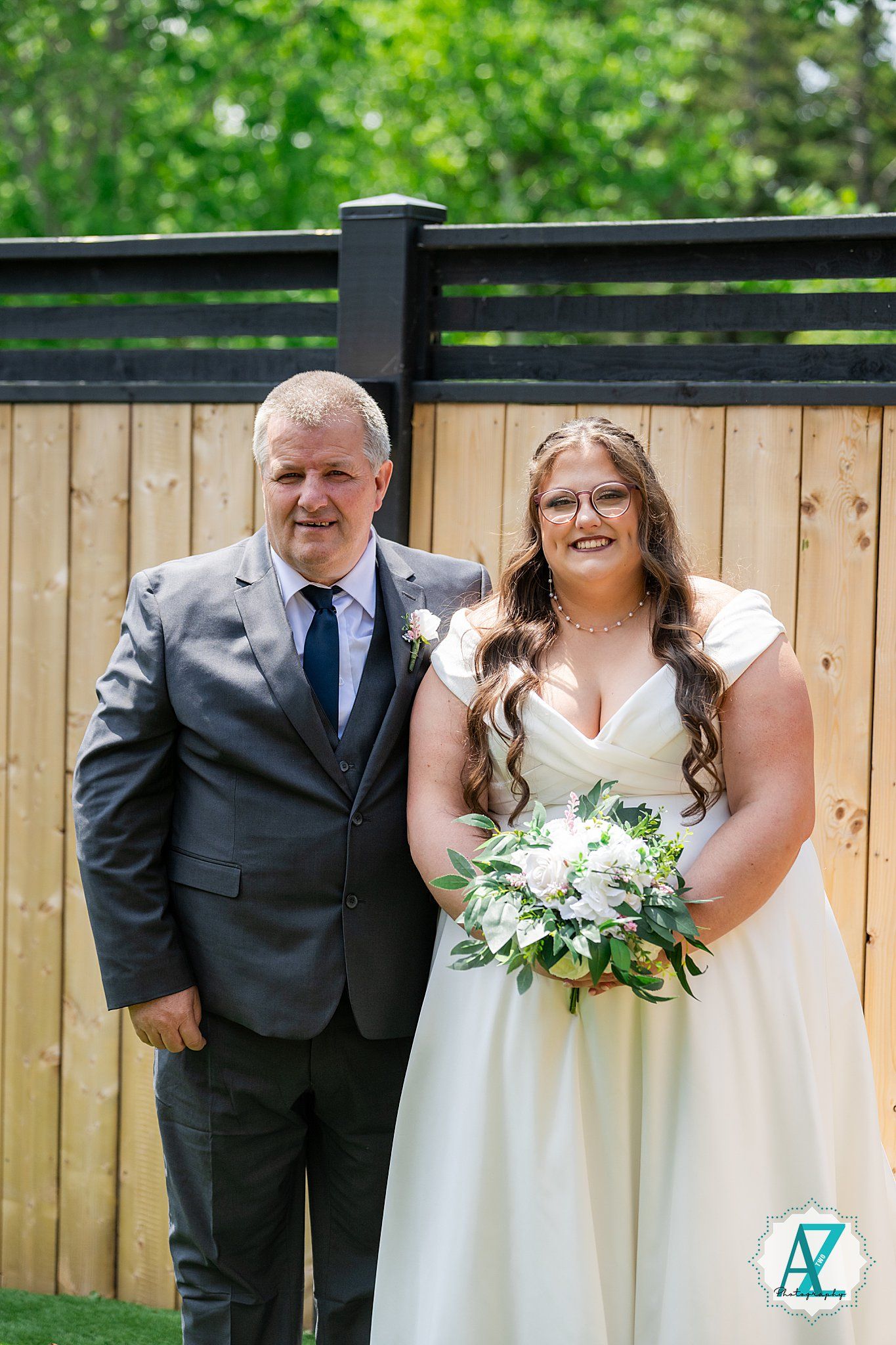 Wedding photo of a couple on their special day standing in front of a wooden fence, with the bride holding a white bouquet.