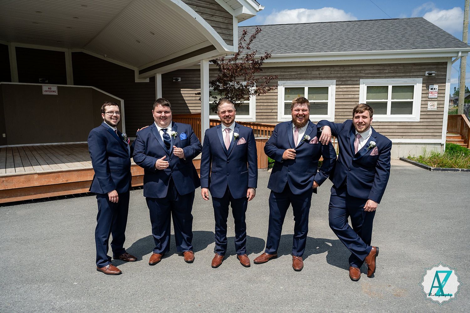Wedding party members in matching navy suits and brown shoes pose together on paved driveway outside ranch-style home.