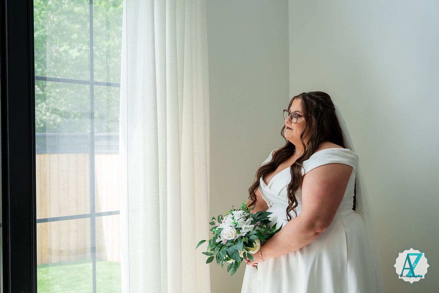 A bride in a white wedding gown holds a green and white bouquet while standing by a sunlit window.