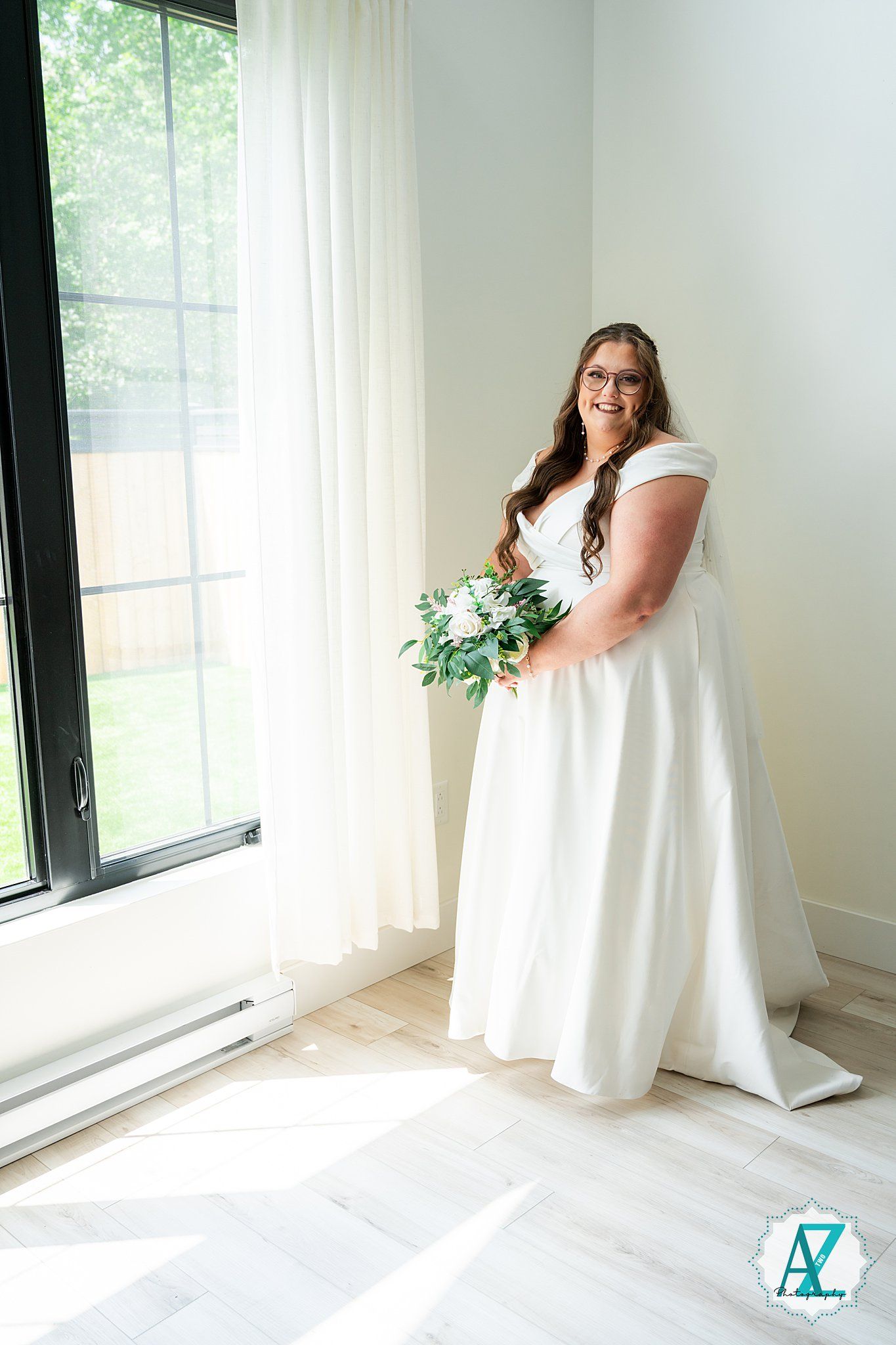 A bride in a white wedding dress stands by bright windows in a minimalist white interior for a series of bridal portraits.
