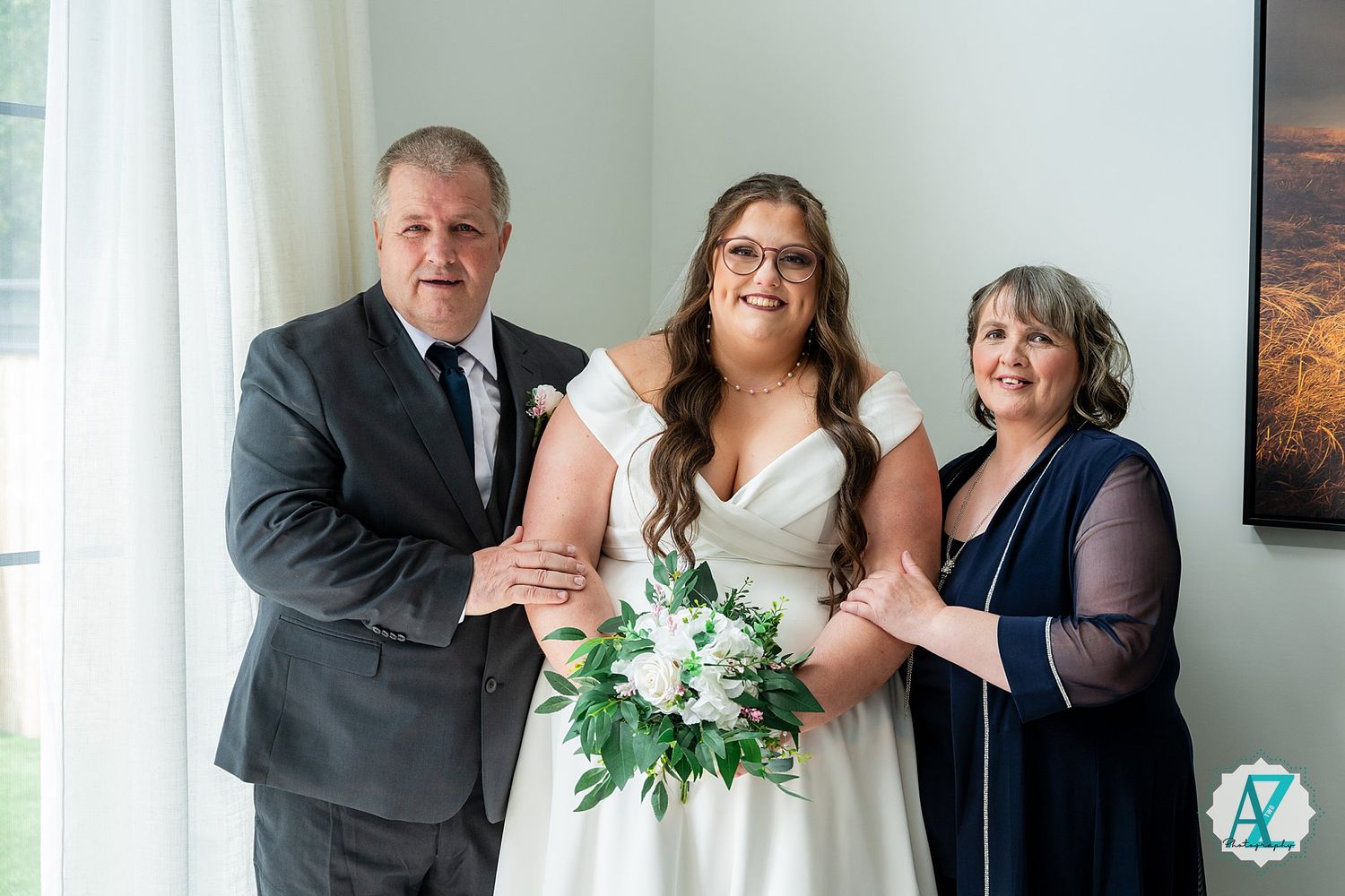 A bride in a white dress holds a green and white bouquet while standing next to two guests at an indoor venue.