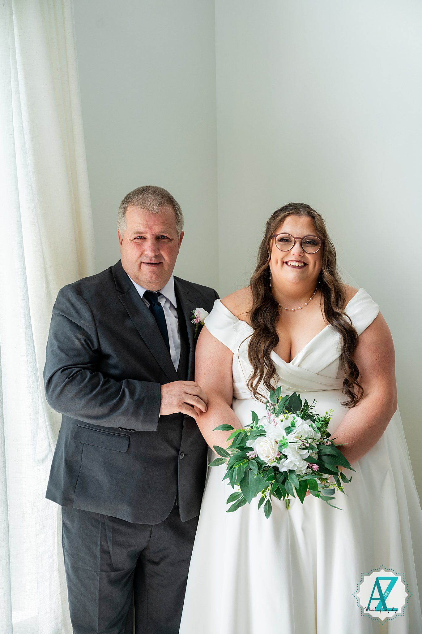 A couple poses together on their wedding day, with the bride in white holding a green and white bouquet.