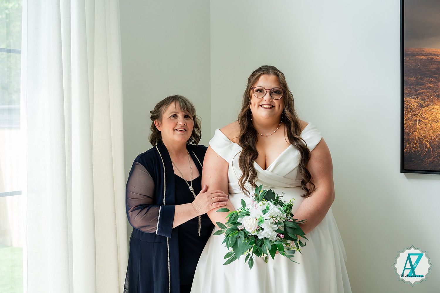 A person in a black mesh top stands with another in a white wedding dress holding a white and green bouquet near a window.