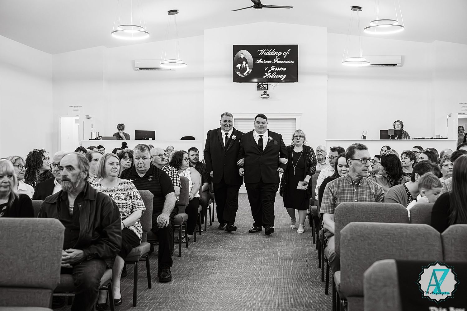 Black and white photo of people seated in rows at an indoor gathering with two figures walking down the center aisle.
