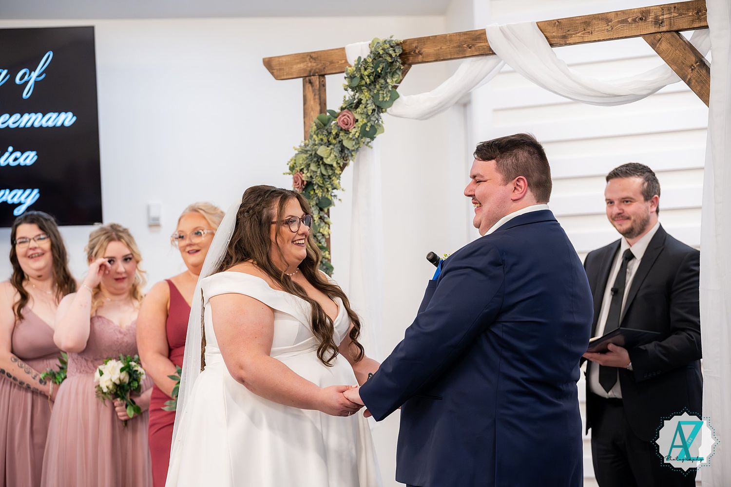A couple exchanging vows under wooden arch decorated with greenery while wedding party looks on.