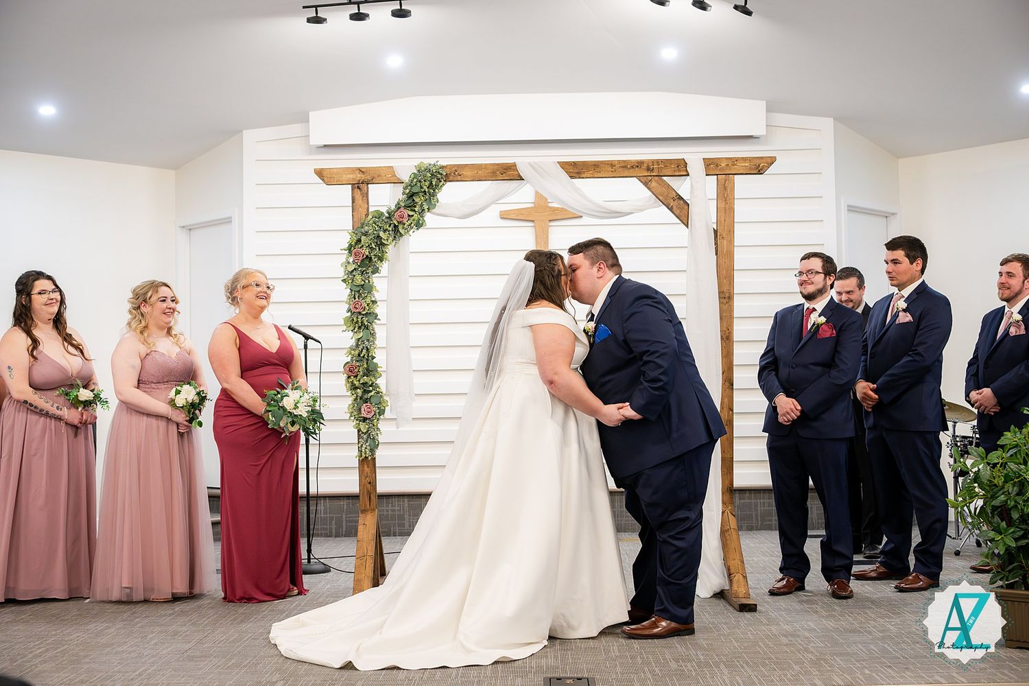 Wedding ceremony taking place under wooden arch decorated with greenery as bridal party watches in pink and navy attire.