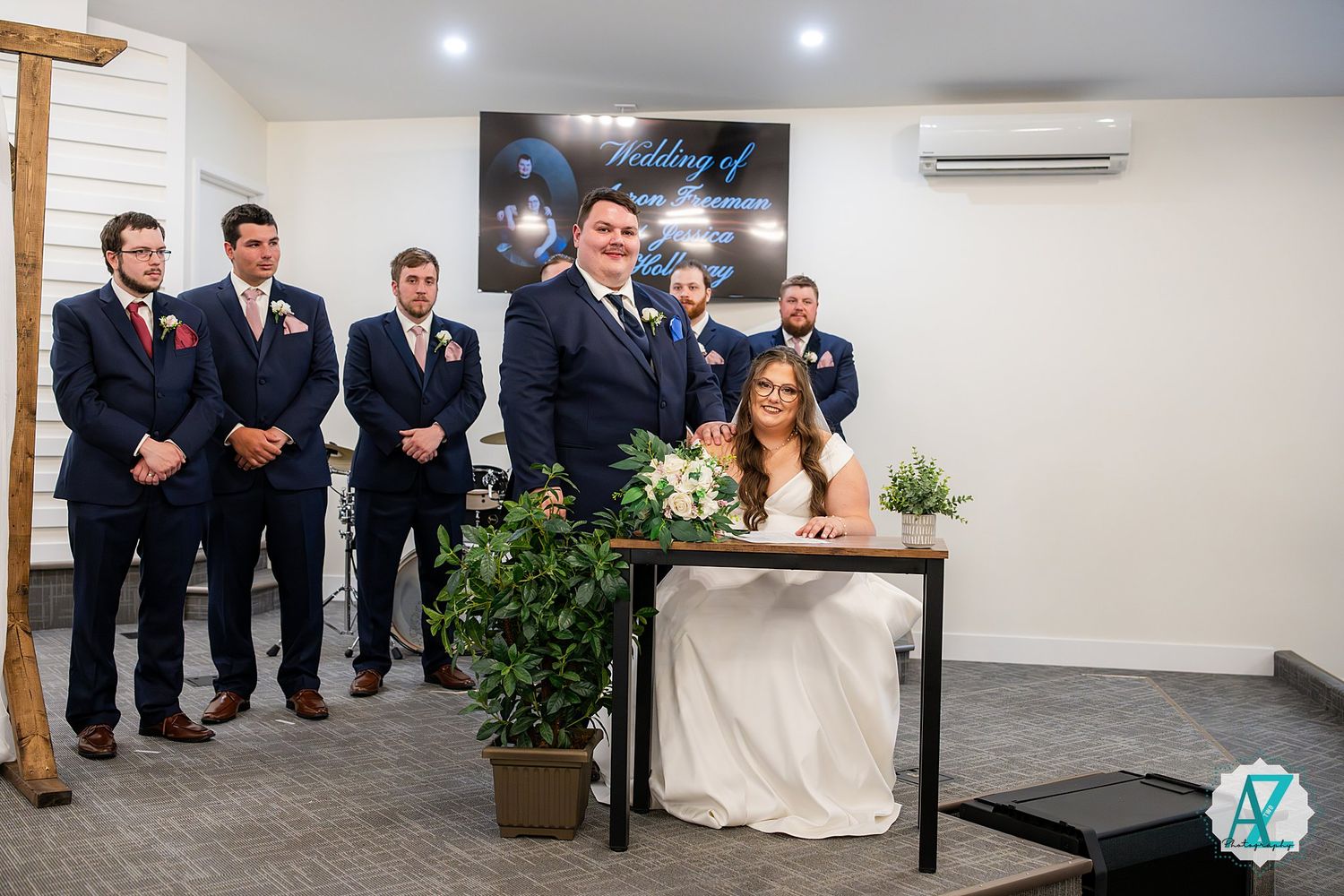 Wedding party stands in formal attire against a white wall with indoor plants and decorative lighting.