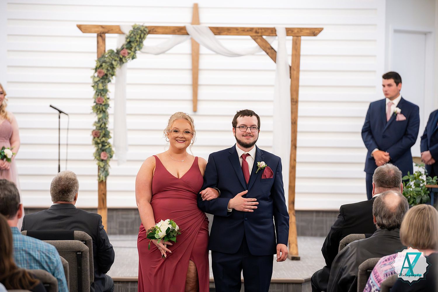 Two people walk arm in arm down the aisle at a wedding ceremony with wooden cross arch and floral decor in background.