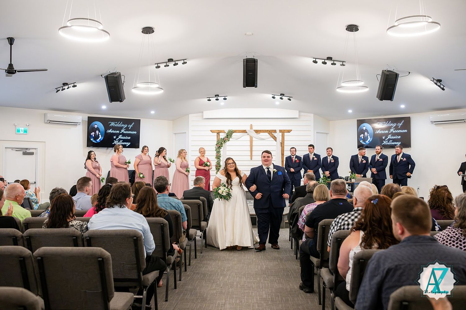 Wedding ceremony inside a bright modern chapel with guests seated in rows, decorative lighting and AV screens mounted on walls.
