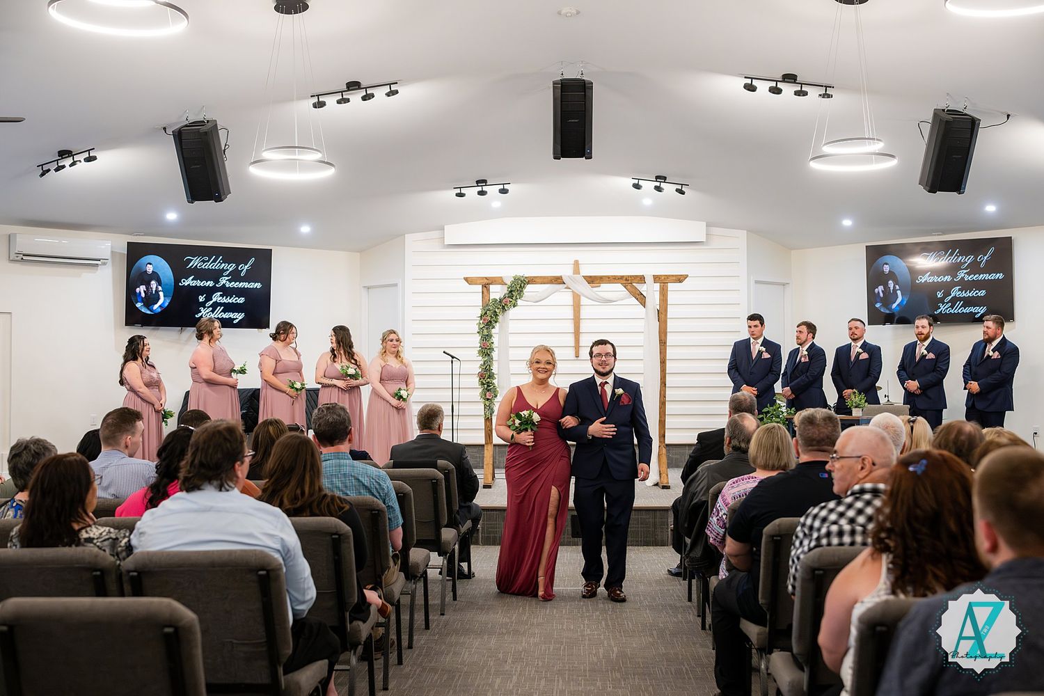 Wedding ceremony in progress at a small indoor chapel with bridesmaids in pink and groomsmen in dark suits standing along sides.