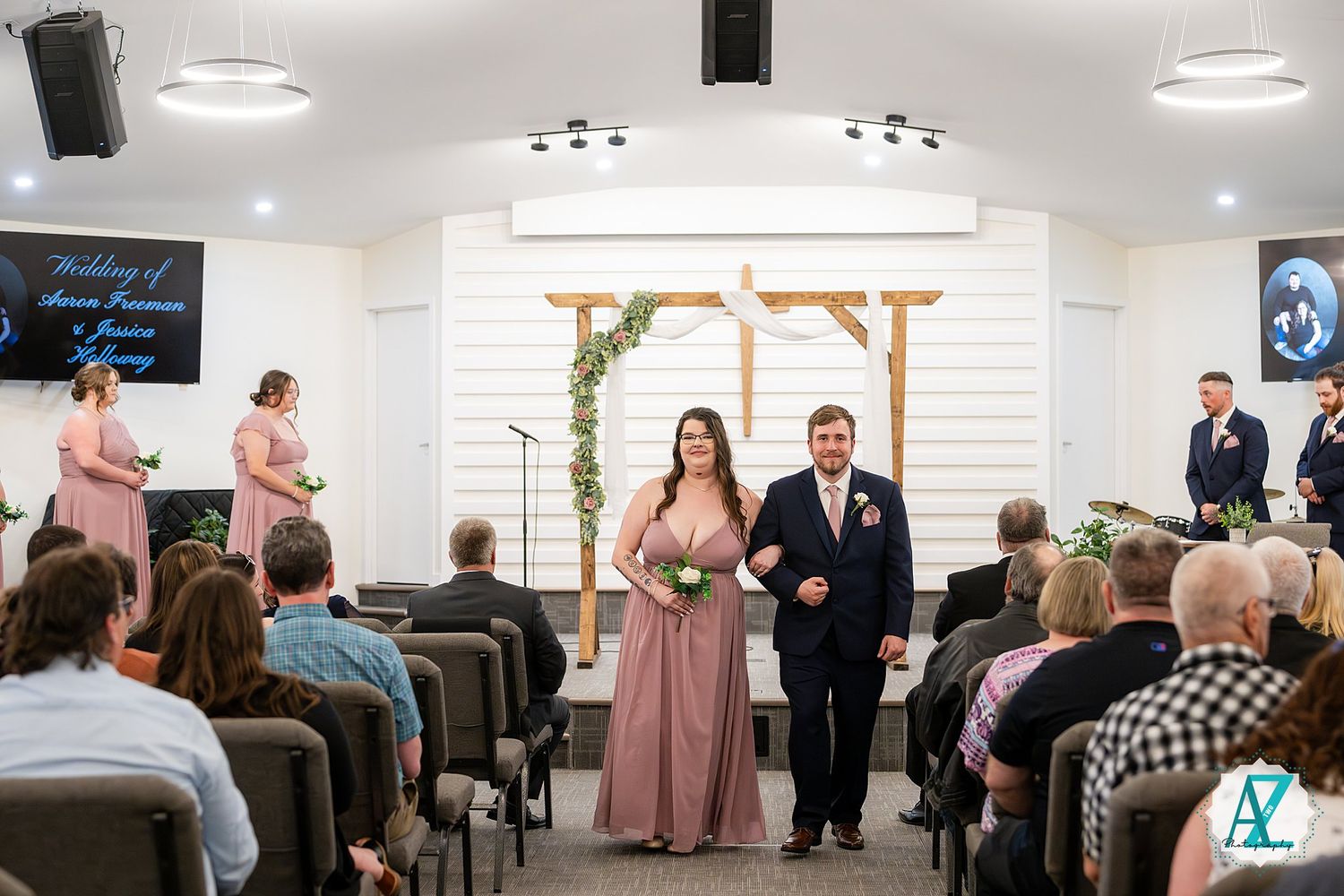 A wedding ceremony in progress inside a modern white chapel with guests seated in rows and attendants walking down the aisle.