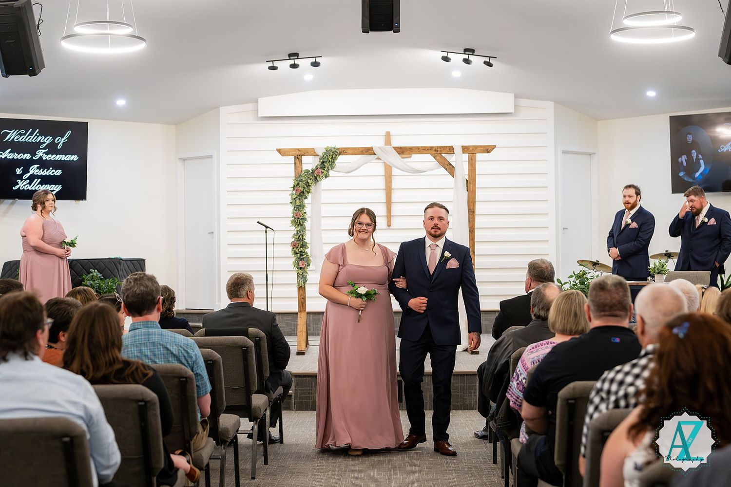 Intimate indoor wedding ceremony with guests seated in rows watching couple exchange vows in front of wooden arch.