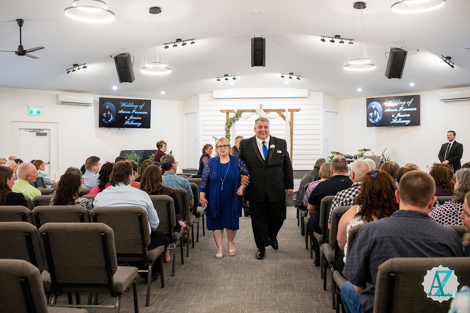 Religious ceremony taking place in modern church sanctuary with pews filled and two people walking down center aisle.
