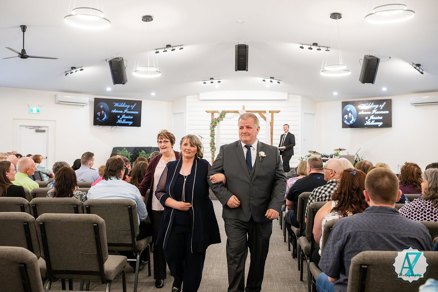 A wedding ceremony processional in a modern church sanctuary with rows of guests seated on gray chairs and TV screens mounted.