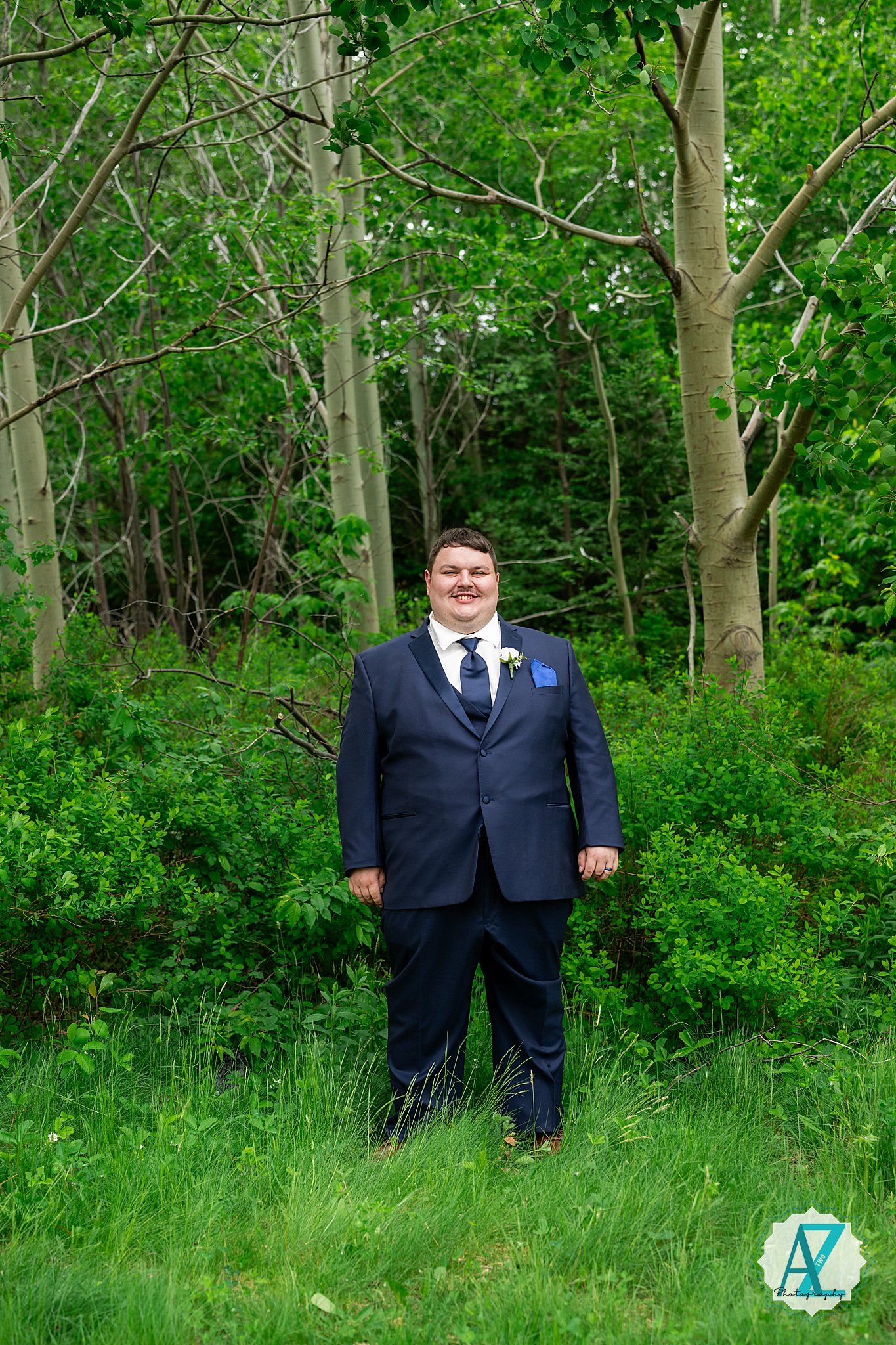 Groomsmen in navy suits stand in a green wooded area during an outdoor wedding photoshoot.